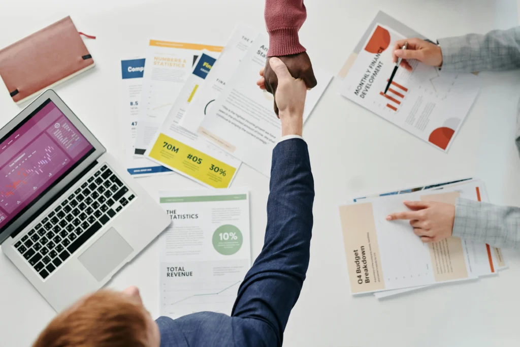 Business professionals shaking hands over marketing reports and analytics dashboards, symbolising a successful digital marketing partnership and business growth.