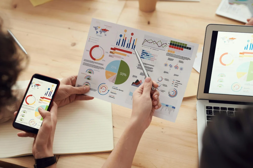 Business team reviewing data analytics dashboards and performance charts on paper, smartphone and laptop to evaluate marketing results and business growth.